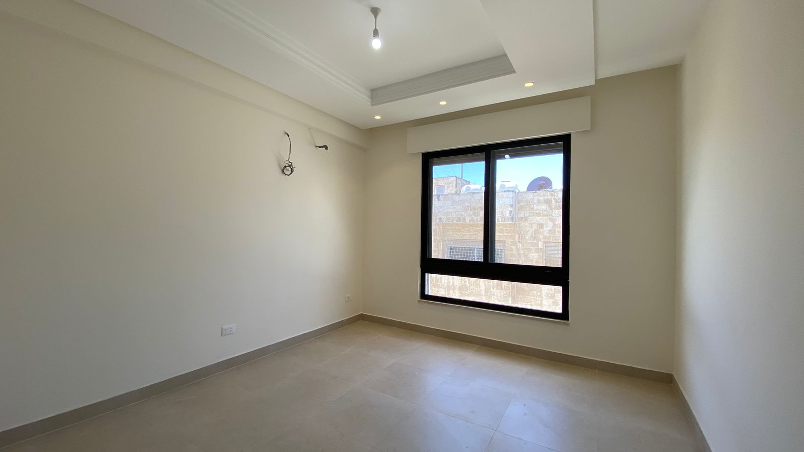 ebb2497f-a0d0-4c3a-b8cd-3b6ab1e4d79d Empty modern room with beige walls, a large window letting in daylight, tiled floor, and exposed wires on the left wall under a simple ceiling light.