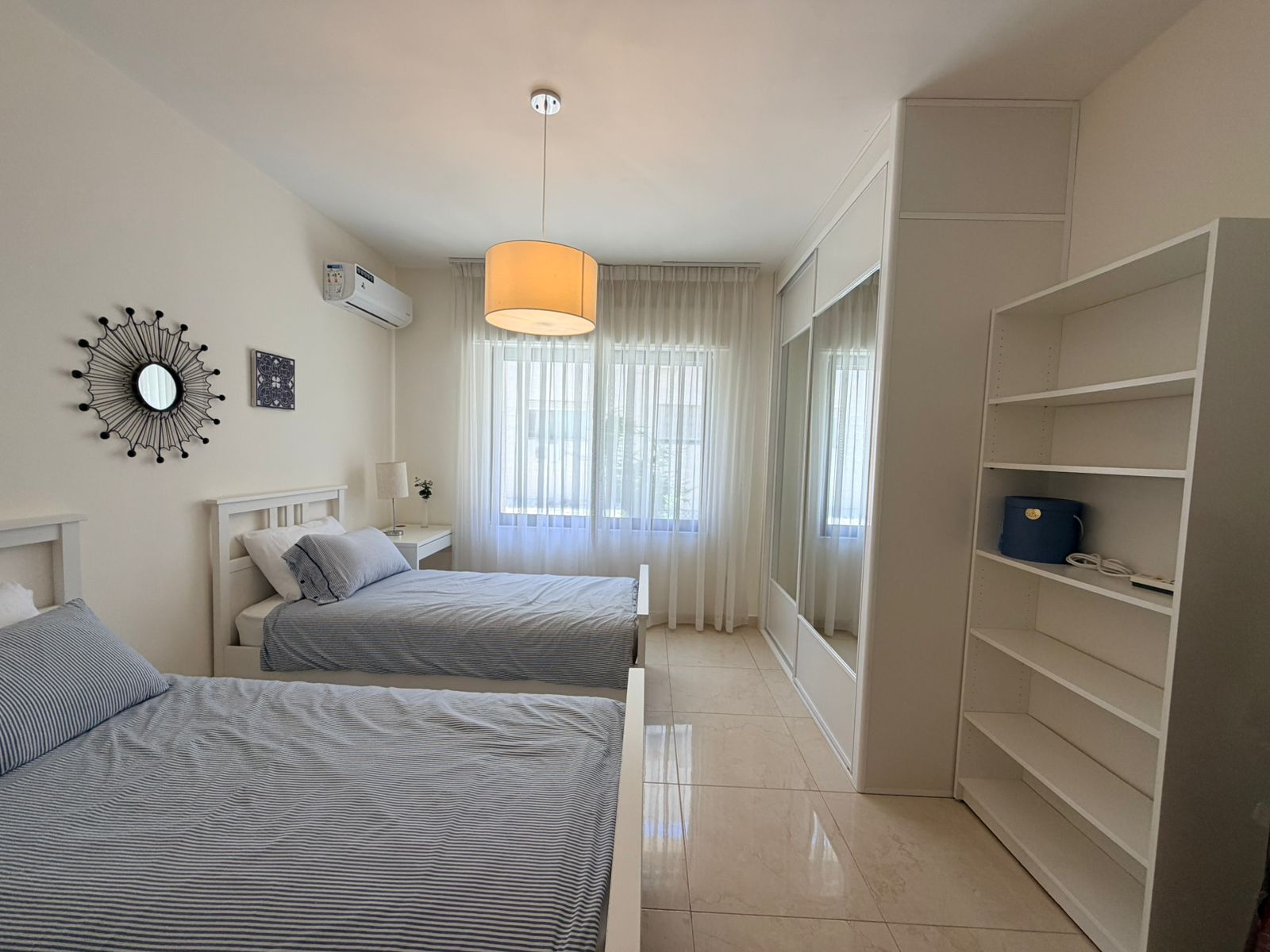 ed239509-ca62-4498-9c7e-17f3a23ff955 Bright white bedroom with two single beds, blue striped bedding, nightstands, and a large mirrored wardrobe.