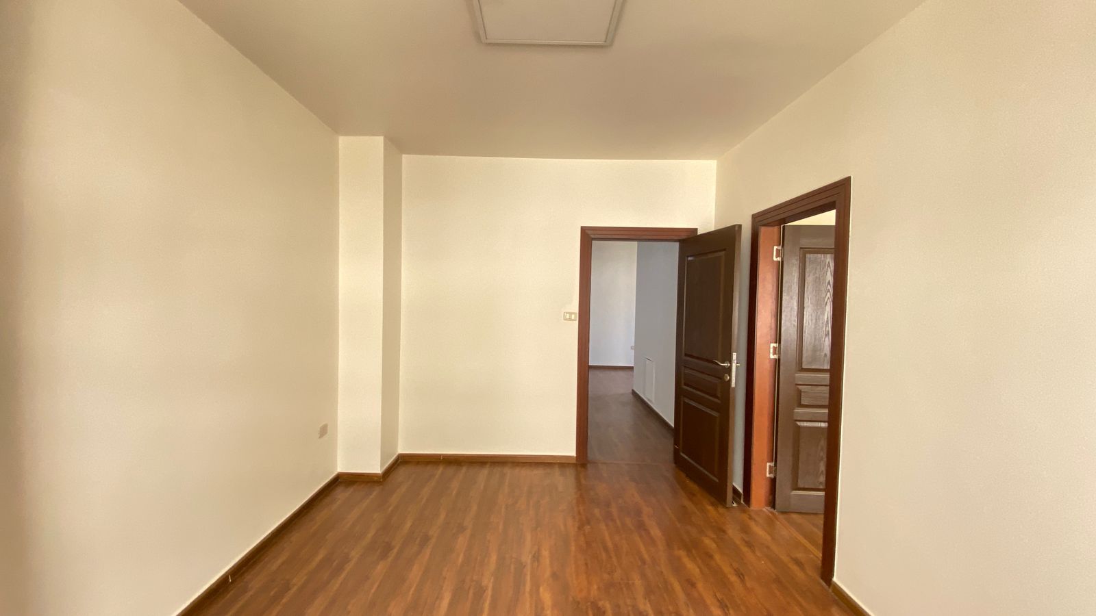 edb16c6a-52f3-490e-94c1-a64fb50665ee Empty beige-walled room with dark wood baseboards and door frames; doorway open to adjacent space.