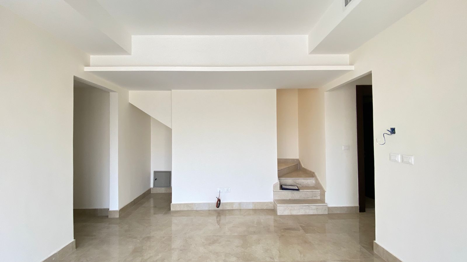 eef9dd38-fd3e-4fe5-ae07-c75e1ba5685e Empty modern living area with beige tiled floor, white walls, and a staircase on the right leading up to a landing beside a central wall