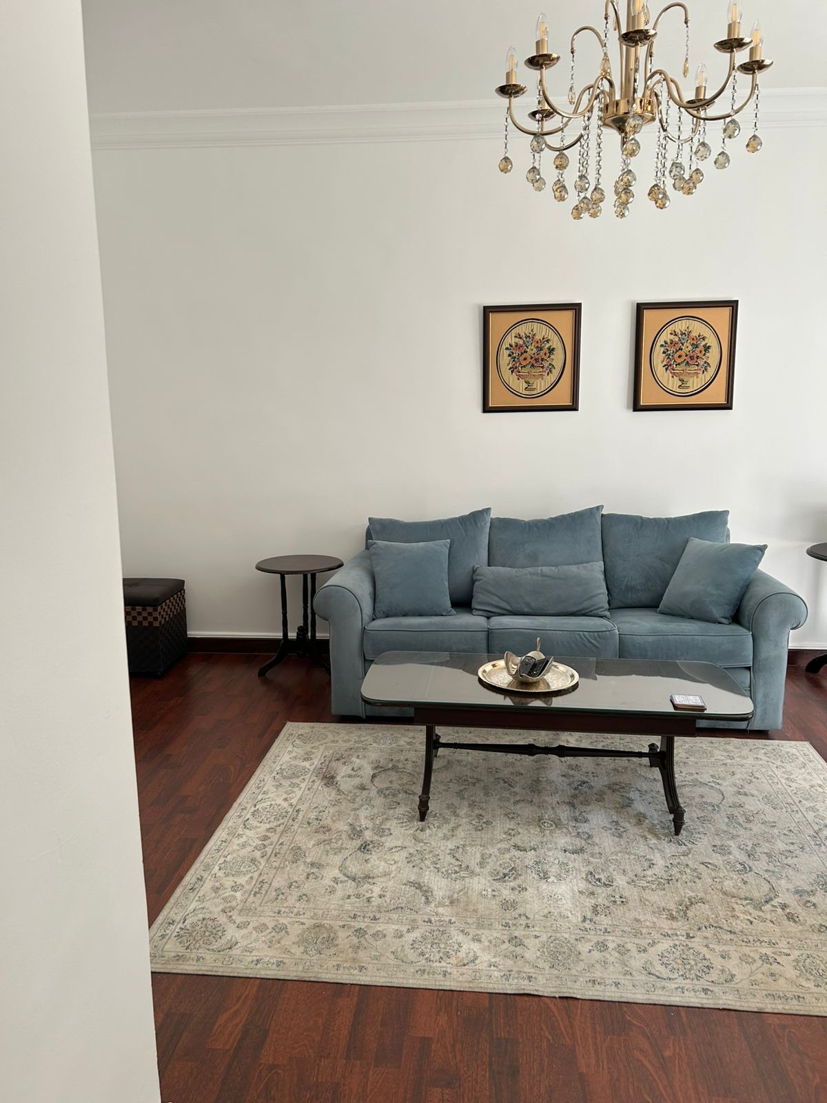 eefcc075-2682-4335-b168-9a7c6a479641 Light blue sofa with matching cushions in a living room, glass coffee table, crystal chandelier overhead, two framed floral artworks on the wall, and a light-patterned rug.