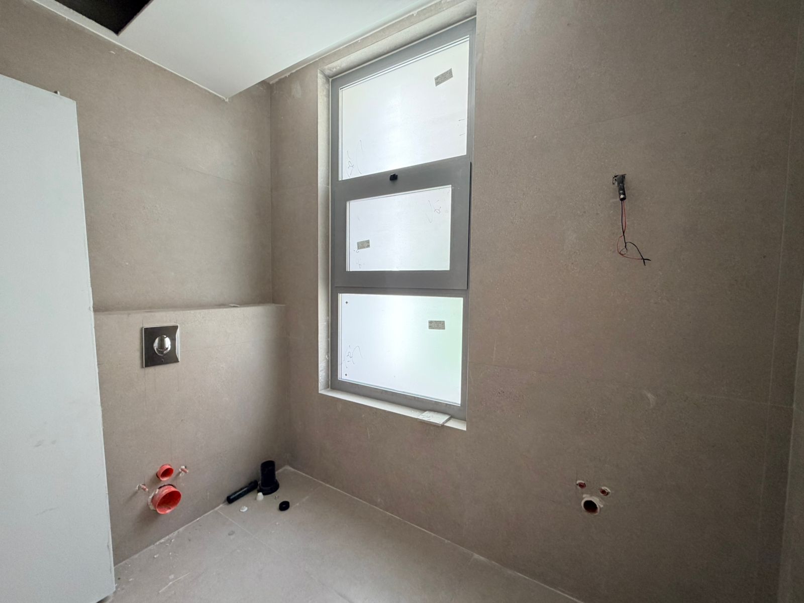 f00fa159-ab78-4510-80ec-9201459b65ce Under-construction bathroom with a large frosted window, exposed red plumbing pipes near the floor, and loose wiring on the wall. Tall white cabinet on left side.