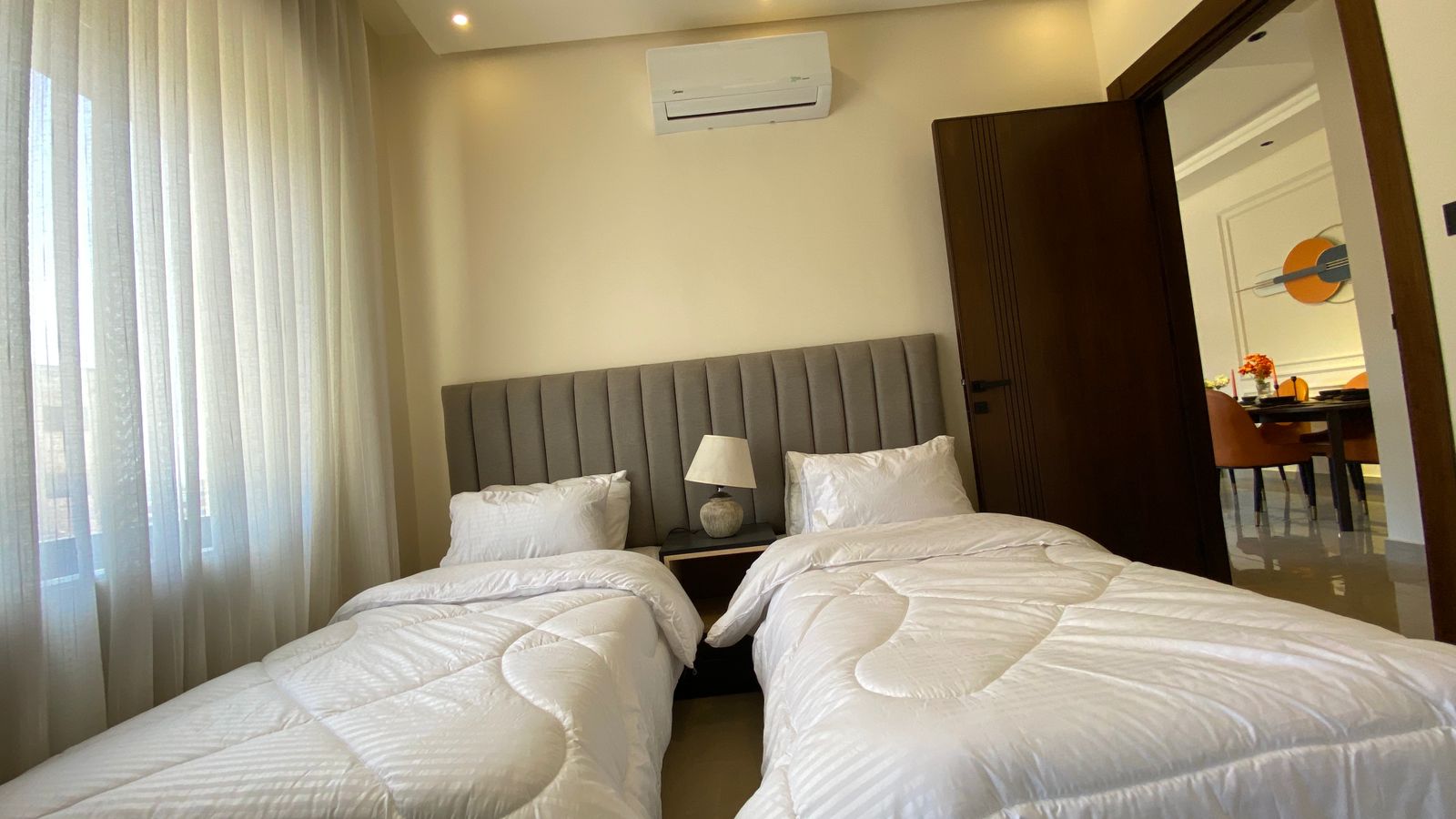 f0b4b448-b3c3-4c01-8b59-34e1ce163e2f Hotel room with two twin beds, gray upholstered headboard, and a shared nightstand with lamp between them.