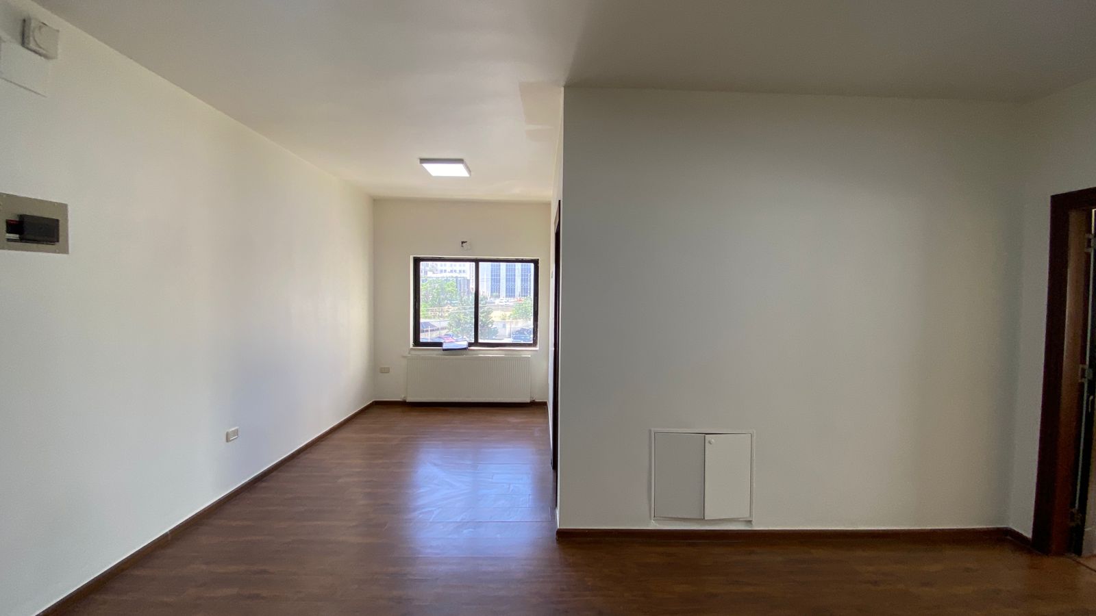 f0e73d24-6c46-4a5c-8076-e4b514fc9489 Empty living room with white walls, wood floors, a large window at the far end, and a radiator beneath it.