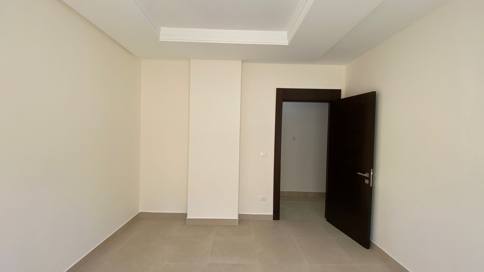 f1238c2a-bf0e-4b04-a36d-23b0bfe555b1 Empty, unfurnished room with light beige walls, tiled floor, and a dark wooden door ajar leading to another space.