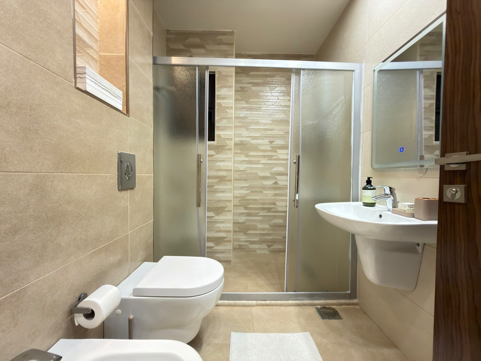 f218bbd9-a32b-4677-8459-5a21724f58b6 Modern beige-tiled bathroom with a frosted-glass shower enclosure, a wall-mounted flush plate, a white toilet, and a pedestal sink with a mirror cabinet nearby.