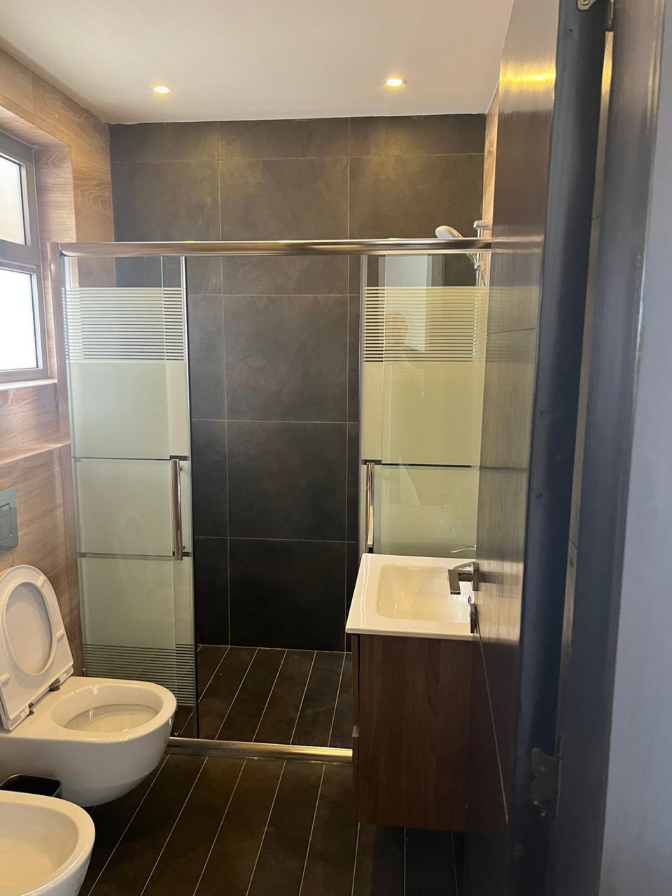 f49404e9-c075-4803-a8bf-d8c74b95dda6 Modern bathroom with dark tile walls and floor, glass shower enclosure, wall-mounted sink, wooden vanity, and toilet with bidet nearby near a window corner.