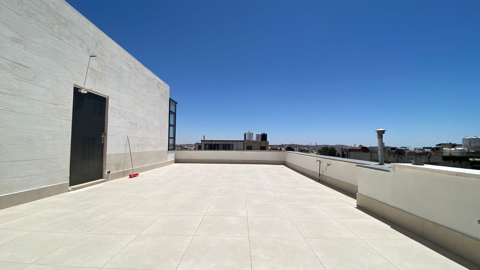 f5c6c880-145f-46f6-8aee-d8049dbc7601 Rooftop terrace with pale tiled floor, a beige wall and door on the left, a broom leaning nearby, and a clear blue sky over distant buildings.