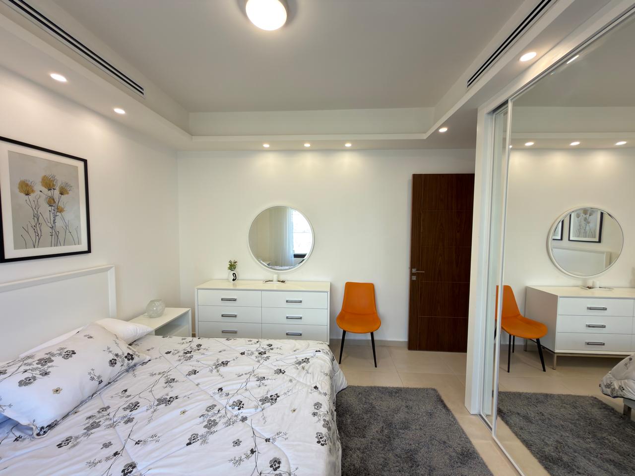 fa0780fd-bfae-4fcb-b6f5-5b94dc24ebc1 Bright modern bedroom with white furniture, round mirror above dresser, orange accent chair, and a mirrored closet door.