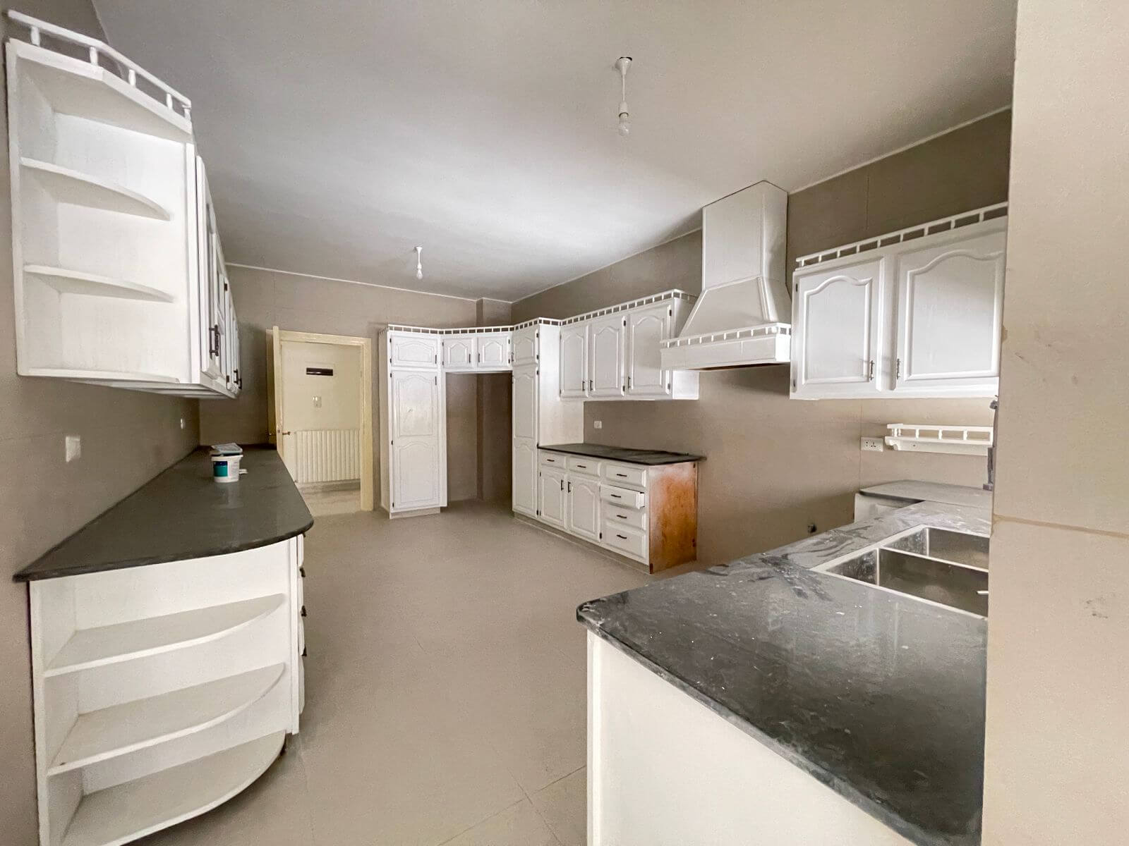 fa08b948-8d3a-4f3f-ba09-fb6a436c8c4a White L-shaped kitchen with tall white cabinets, dark countertops, and a large range hood along the back wall; empty and under-furnishing.