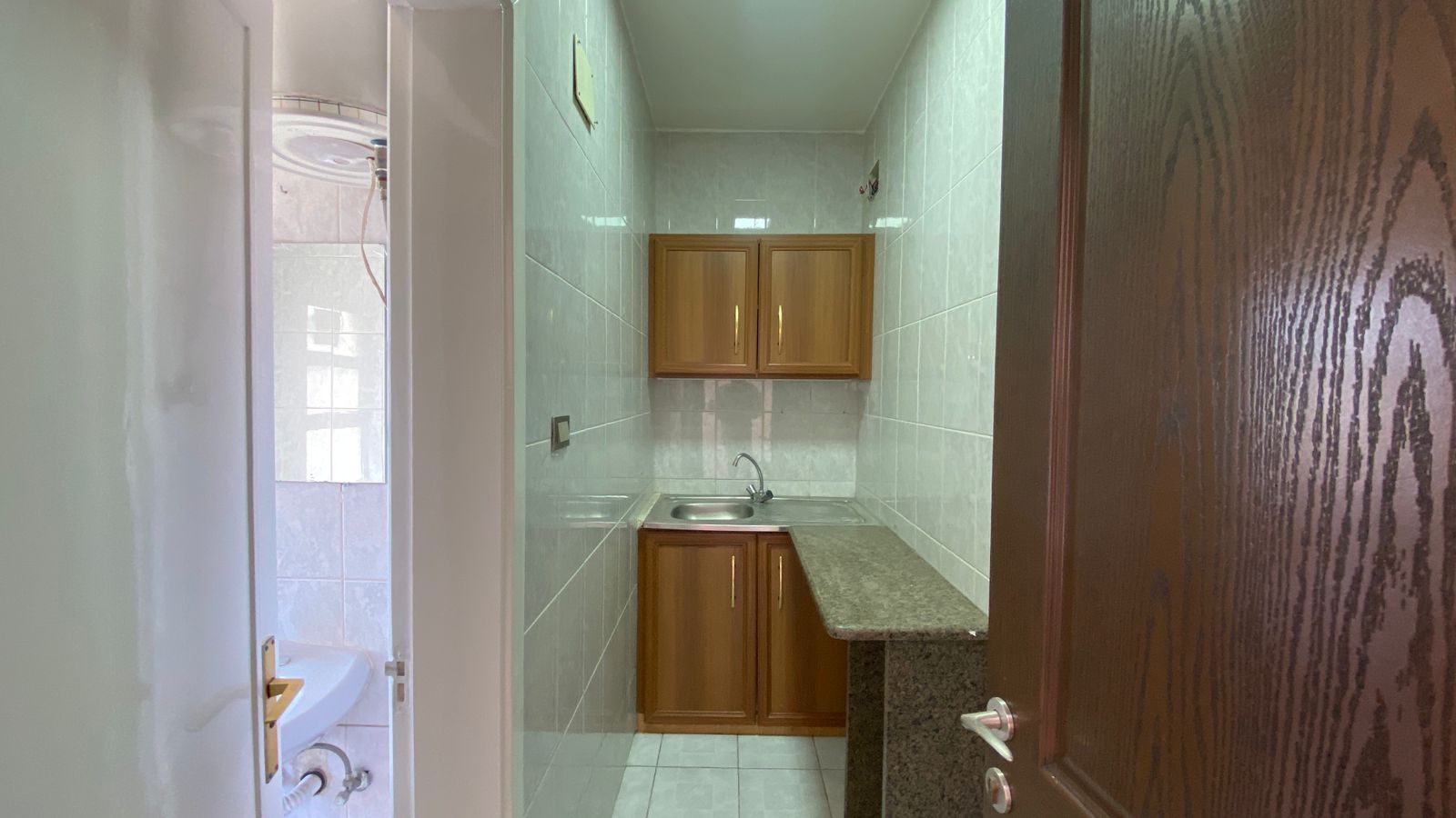 fa1a06e6-4f24-475c-82d9-6de4bbf70b06 Narrow kitchen with green tiled walls, wooden upper and lower cabinets, and a stainless steel sink under a windowed wall; granite counter extends along the right side.