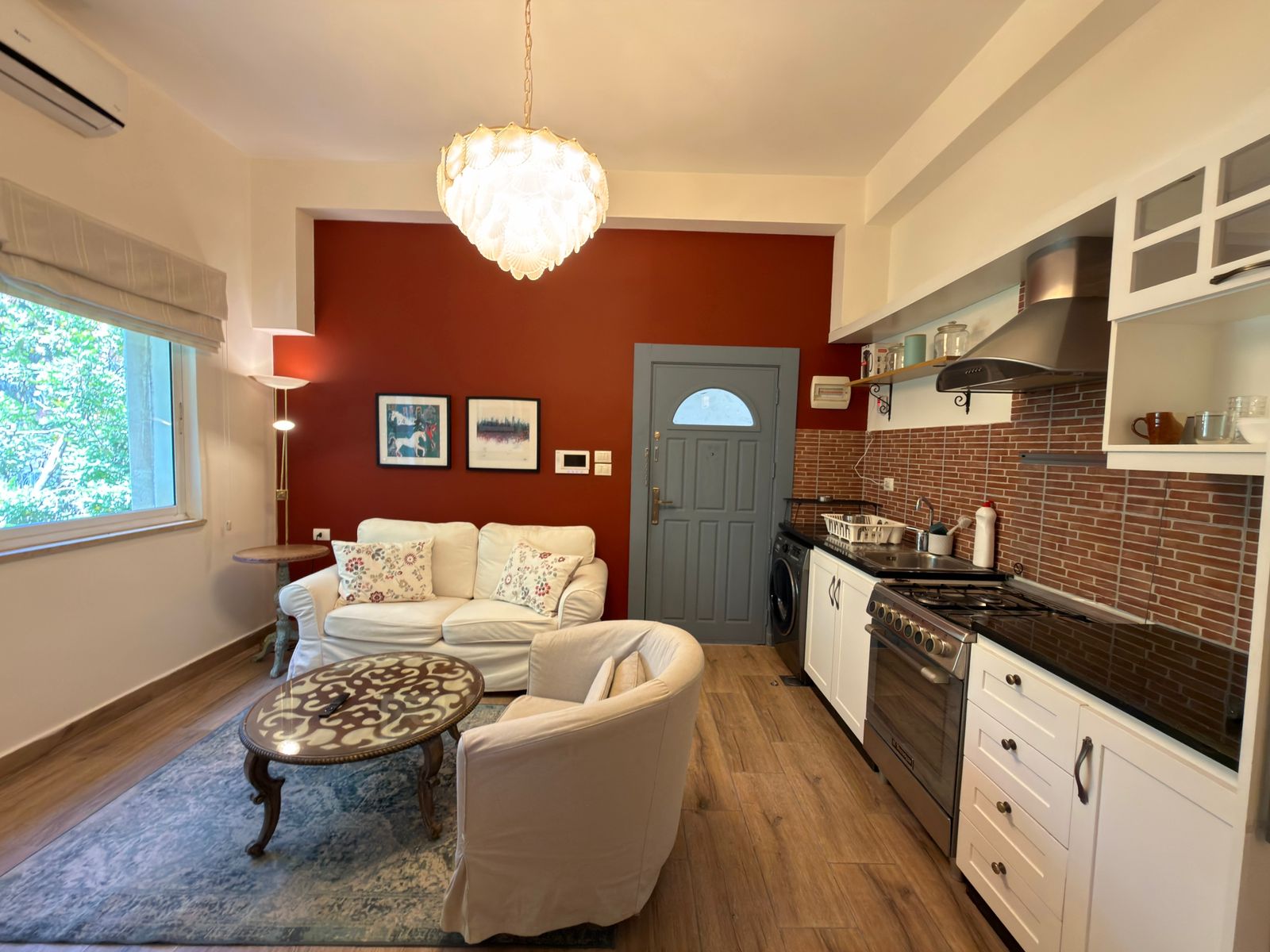 fad52e94-6fa8-4f1b-997c-41428cac2281 Cozy open-plan living room and kitchen with a red accent wall, white sofa, and a decorative chandelier above.