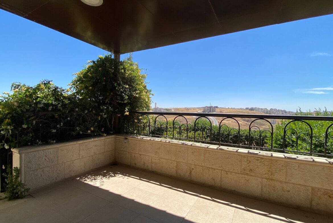 fb3042d5-c8d5-407f-aa0f-97231b8fb56c Balcony with stone walls and a vine-covered corner, overlooking a dry hillside and distant buildings under a clear blue sky.