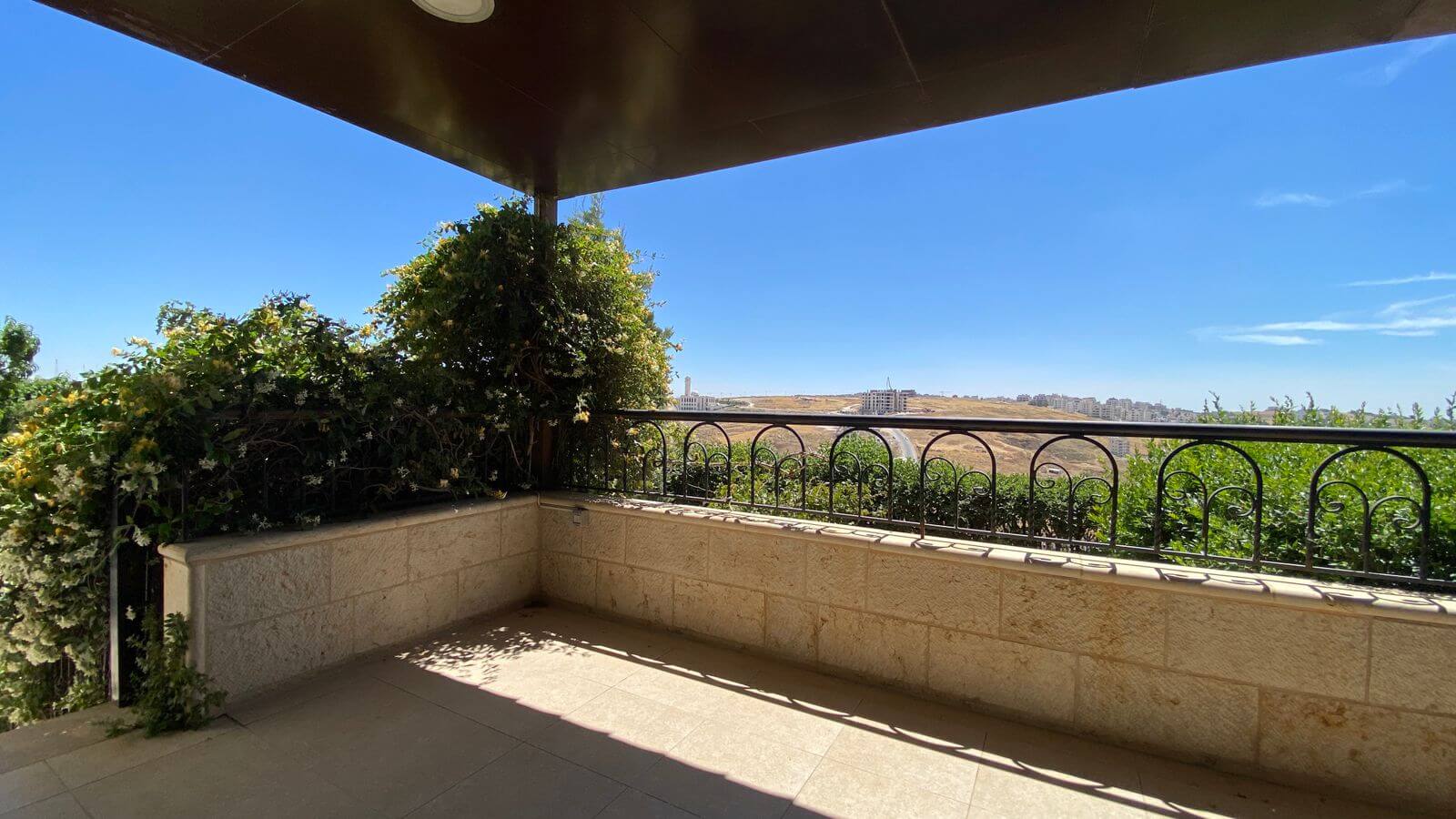 fb3042d5-c8d5-407f-aa0f-97231b8fb56c Balcony with stone walls and a vine-covered corner, overlooking a dry hillside and distant buildings under a clear blue sky.