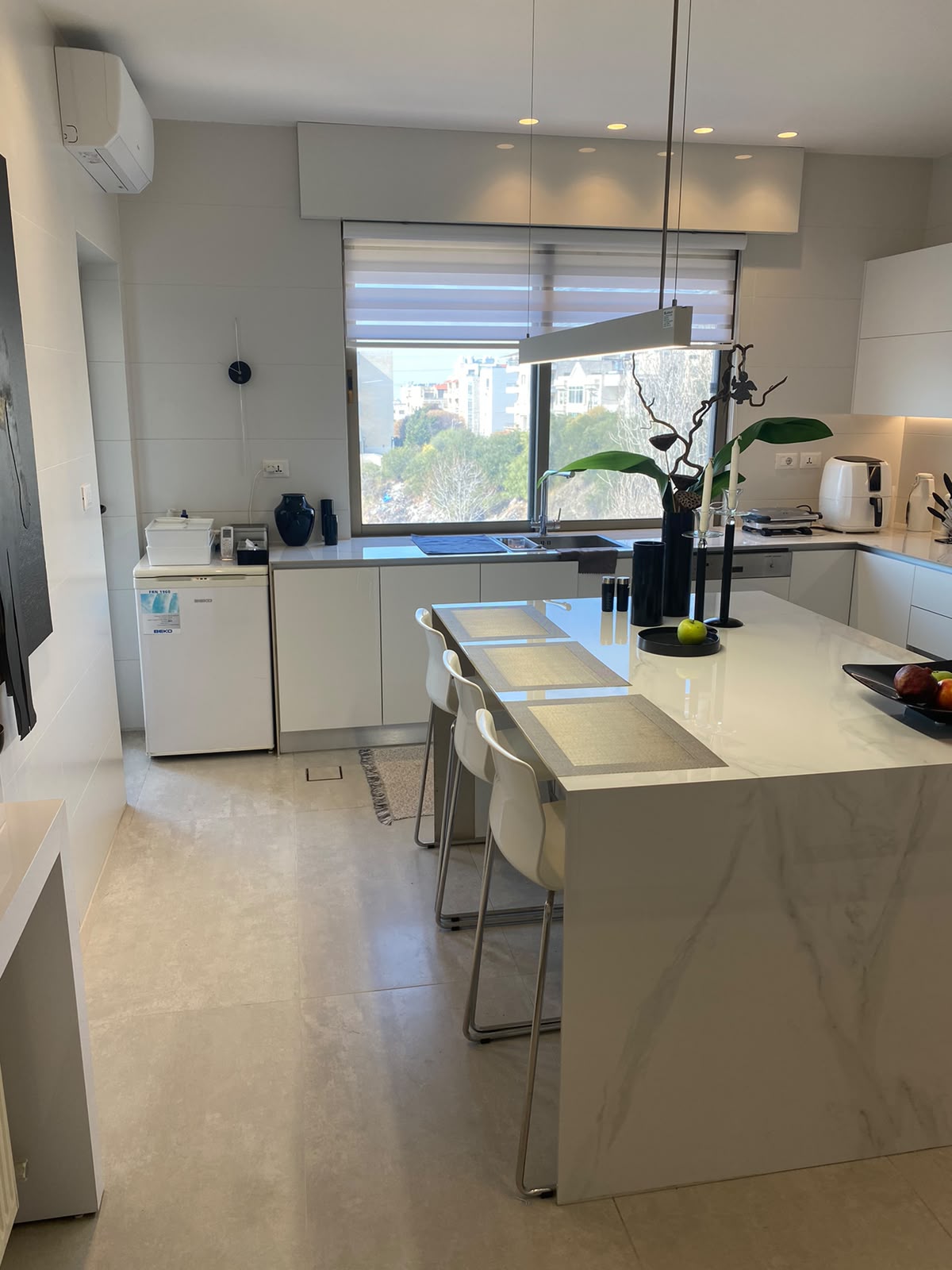 fb4e83ec-e549-4815-971a-61d1df0917c6 Bright modern kitchen with white cabinets, marble island, four white stools, and a city view through a large window.