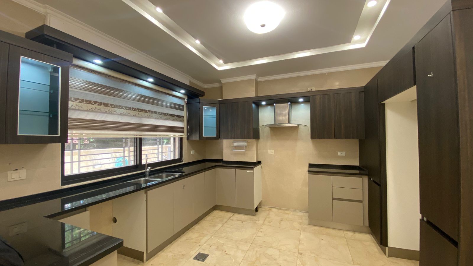 fbb70d60-f743-4e29-a9e0-94927d7b7fbb Modern L-shaped kitchen with dark wood cabinets, black countertops, and beige walls; large window with blinds, double sink, and stainless range hood as centerpiece.