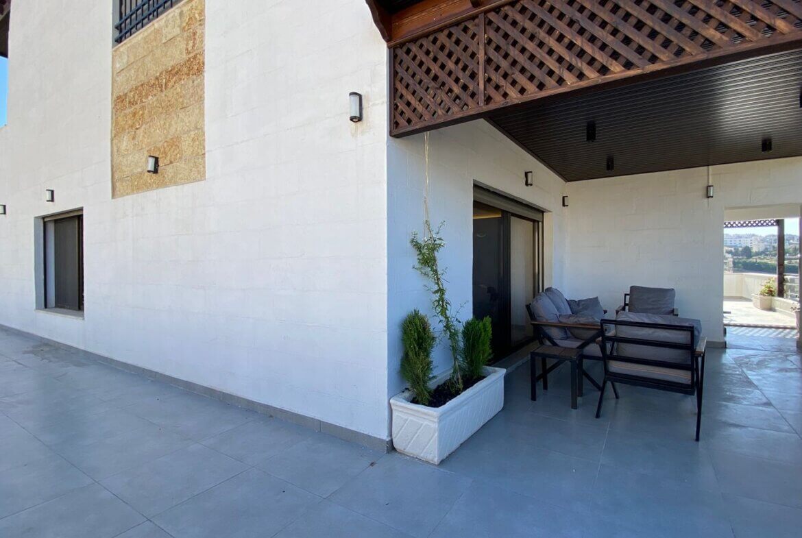 Outdoor patio on a white-walled building with a covered seating area, tiles, and a rectangular planter with small evergreen shrubs.
