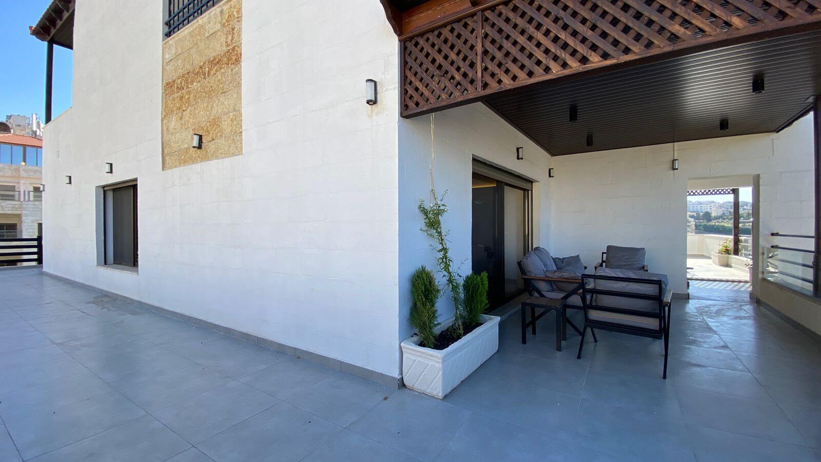 Outdoor patio on a white-walled building with a covered seating area, tiles, and a rectangular planter with small evergreen shrubs.