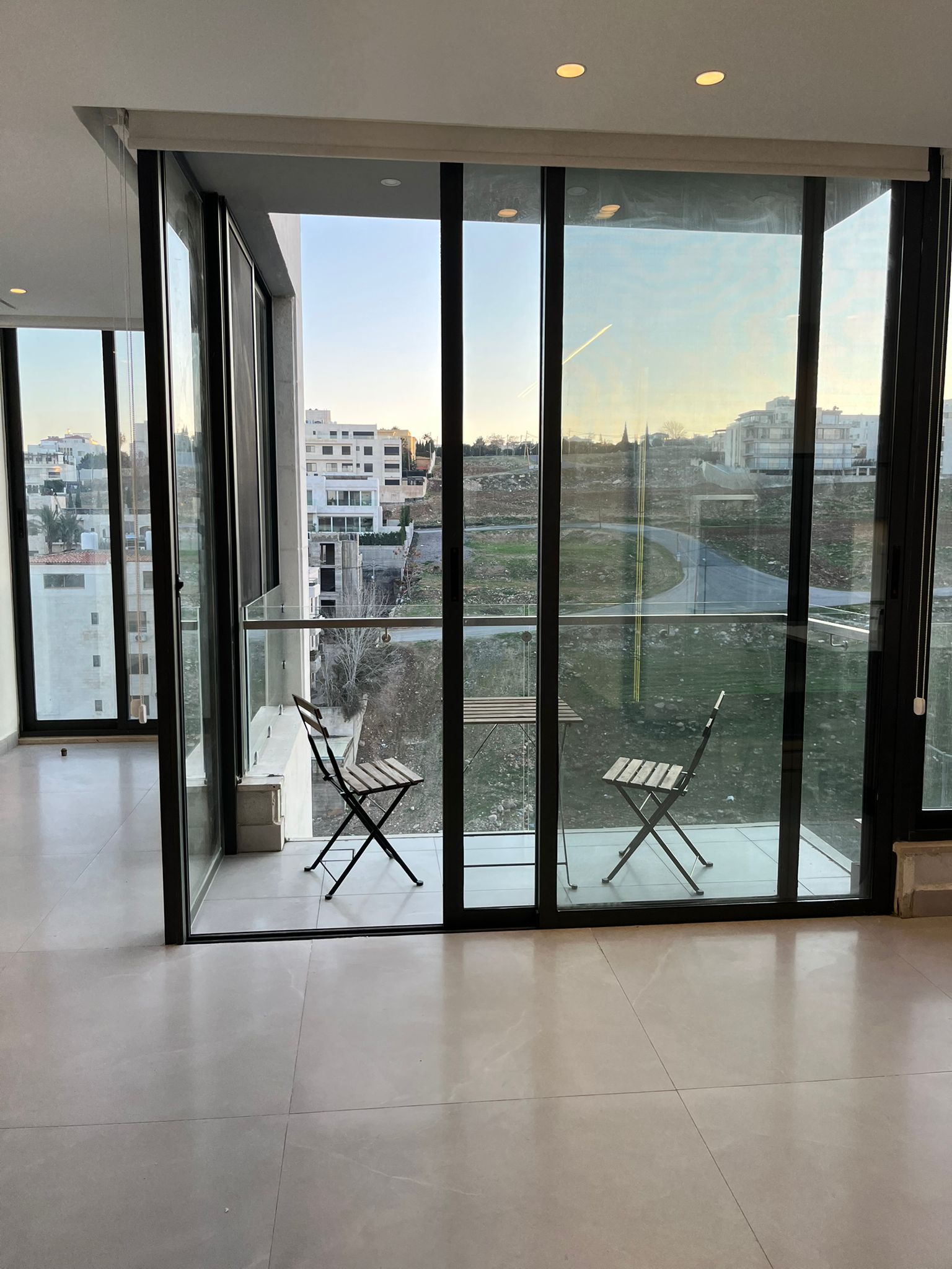 fc5e8836-60a8-4233-bb0c-ef34df2b1a9c Glass sliding doors open to a small balcony with two metal folding chairs; city buildings and an empty lot visible outside.