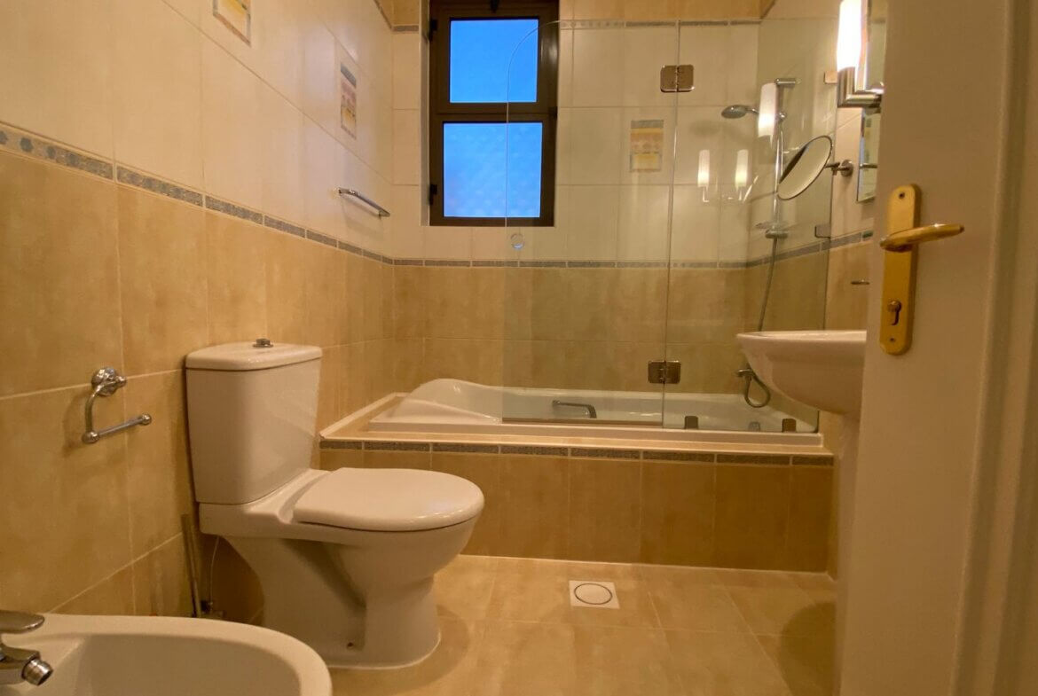 fcc38edb-d436-43c2-97a3-9e25bbea06c2 Cream tiled bathroom with a white toilet and bidet, a glass-enclosed shower over a bathtub, and a small window above the tub.
