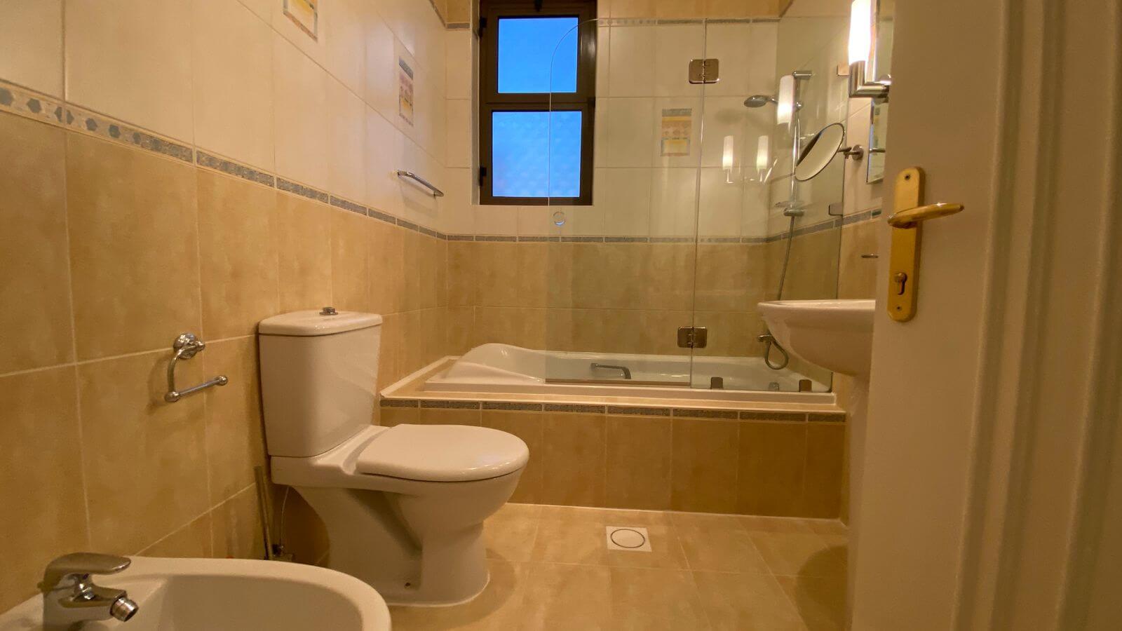fcc38edb-d436-43c2-97a3-9e25bbea06c2 Cream tiled bathroom with a white toilet and bidet, a glass-enclosed shower over a bathtub, and a small window above the tub.