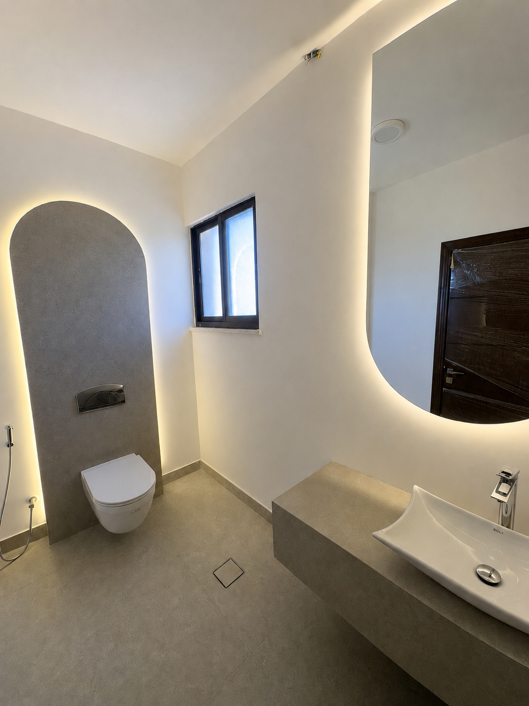 fdac4af5-7e53-4e87-b787-a0f4ee526303 Minimalist bathroom with a wall-mounted toilet, grey curved panel, and backlit edges beside a windowed wall and curved mirror above a concrete countertop sink.