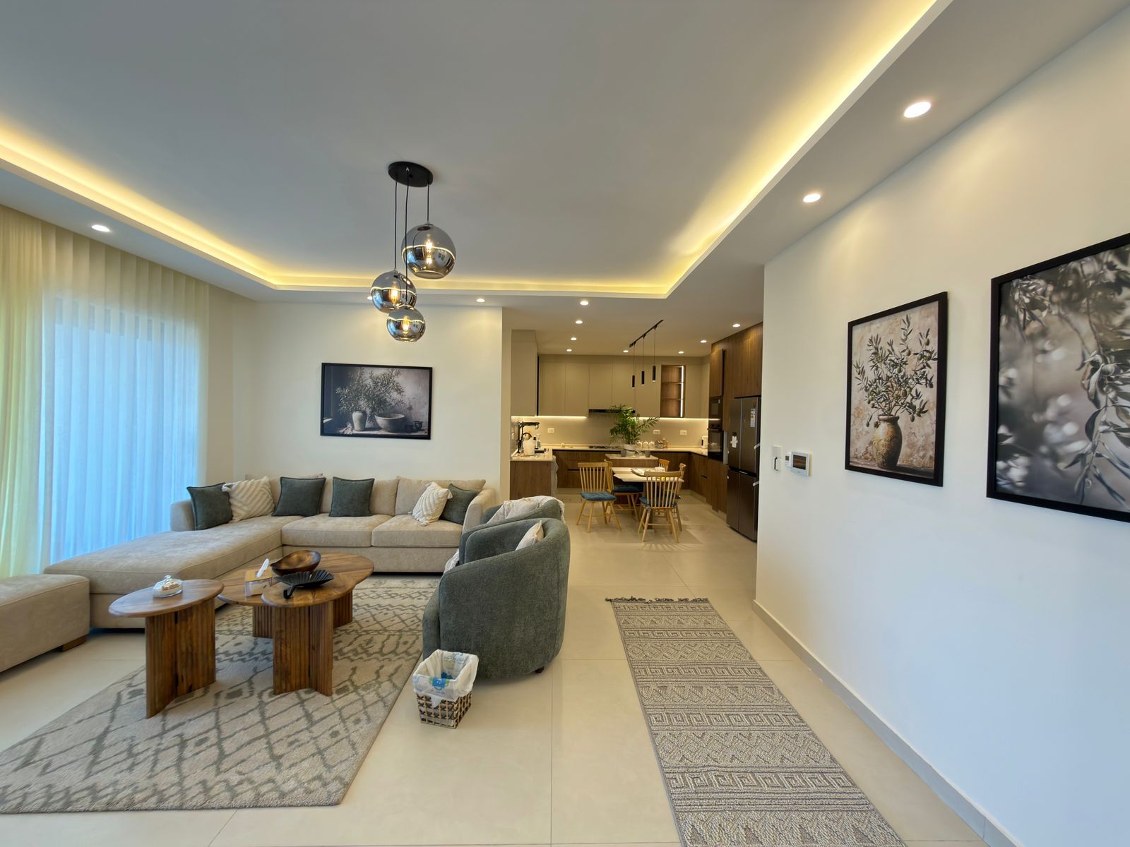 fdea2b4e-5bc6-4b2b-ab40-9dc9a5a63c99 Open-concept living room with a beige sectional, gray chairs, and round wooden coffee tables, opening to a modern kitchen in back.