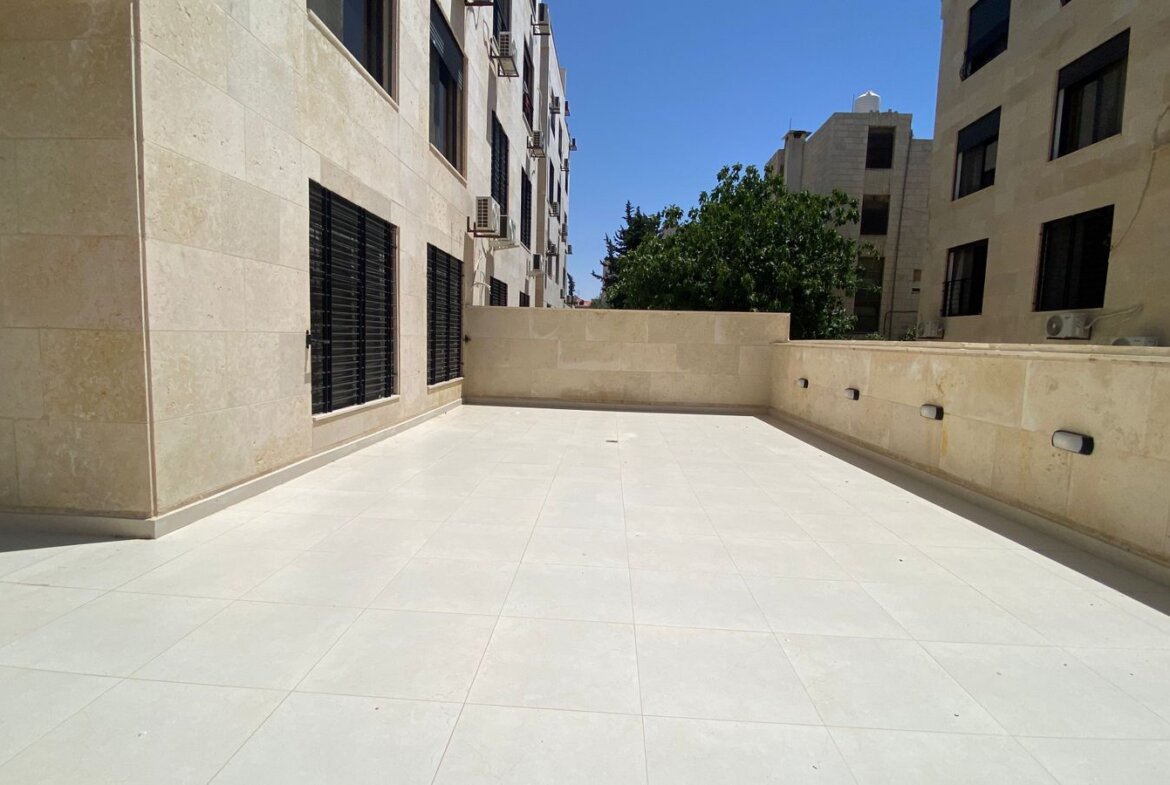 Sunlit rooftop terrace with beige stone walls and light tiles, bordered by a low wall and windows with black bars on the left, air conditioning units mounted on the walls, and a distant tree against a blue sky.