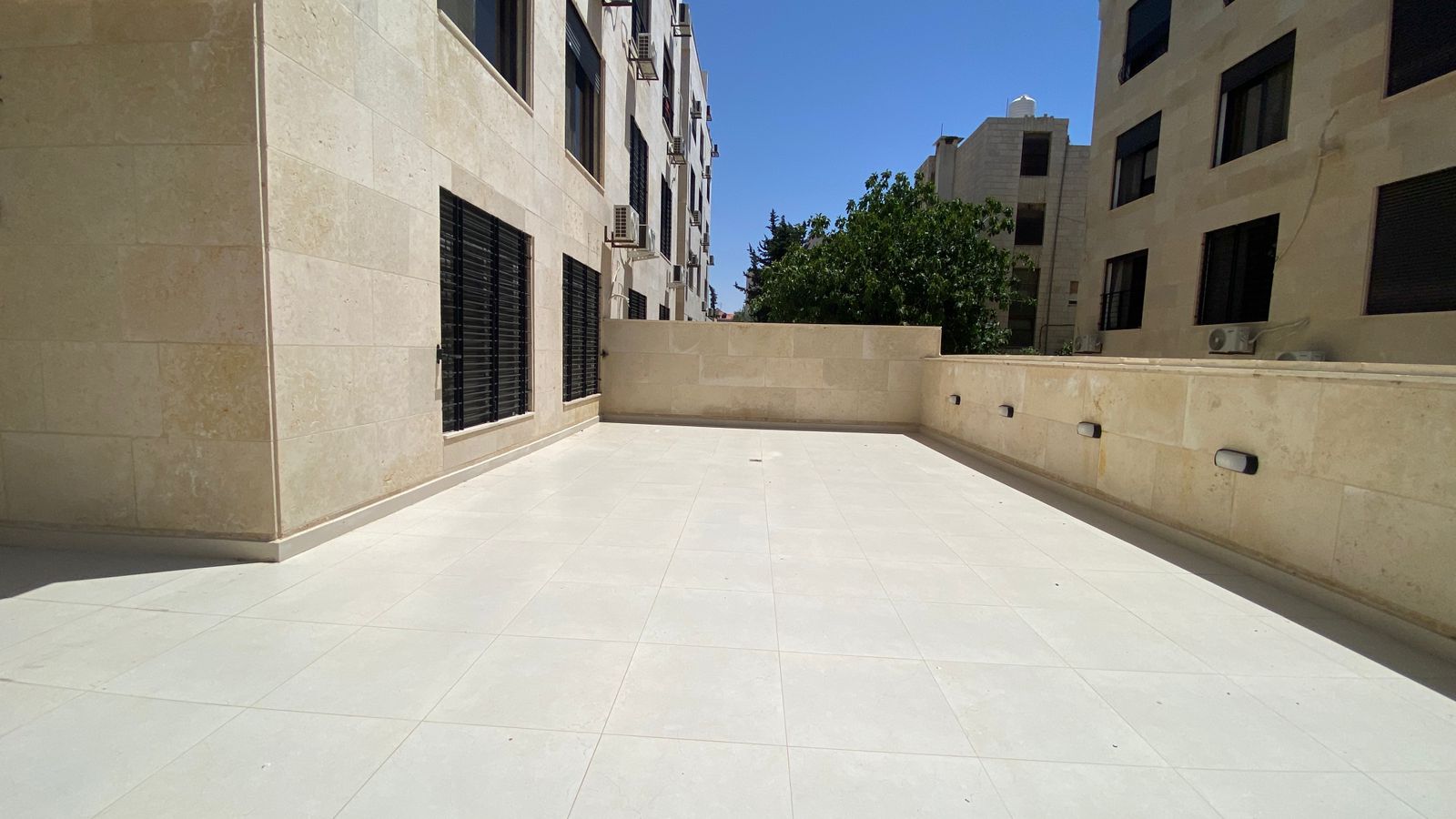 ff77963d-847a-44b9-89d4-2abd228fb629 Sunlit rooftop terrace with beige stone walls and light tiles, bordered by a low wall and windows with black bars on the left, air conditioning units mounted on the walls, and a distant tree against a blue sky.